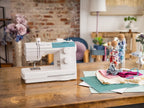 A white sewing machine with a teal accent panel sits on a rustic wooden table in a cozy, softly lit craft room. Surrounding the machine are sewing supplies, including fabric pieces, spools of thread in glass jars, and a wooden spool holder.