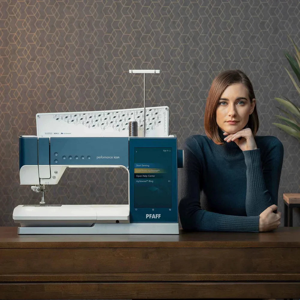Woman sitting next to a PFAFF sewing machine on a wooden table with a patterned wall background