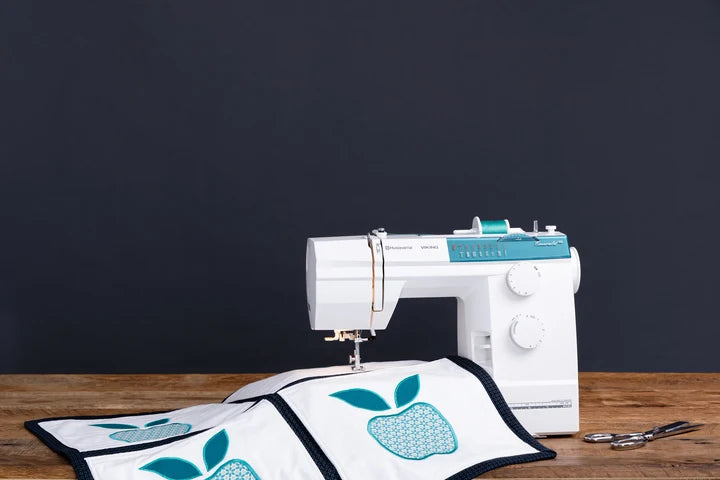 A white sewing machine with teal accents sits on a wooden table next to a white quilted fabric piece featuring a teal apple design, with a pair of scissors on the right, against a dark charcoal background.