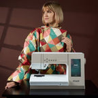Woman wearing a colorful apron sitting next to a PFAFF sewing machine on a dark surface with a brown background.