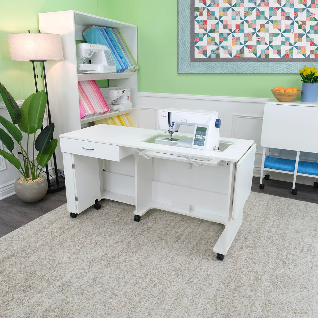 Arrow Christa Sewing Cabinet in White displayed in a sewing room setup with machine installed and quilt hanging on the wall.