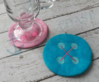 Pink and turquoise embroidered button coasters shown with a glass