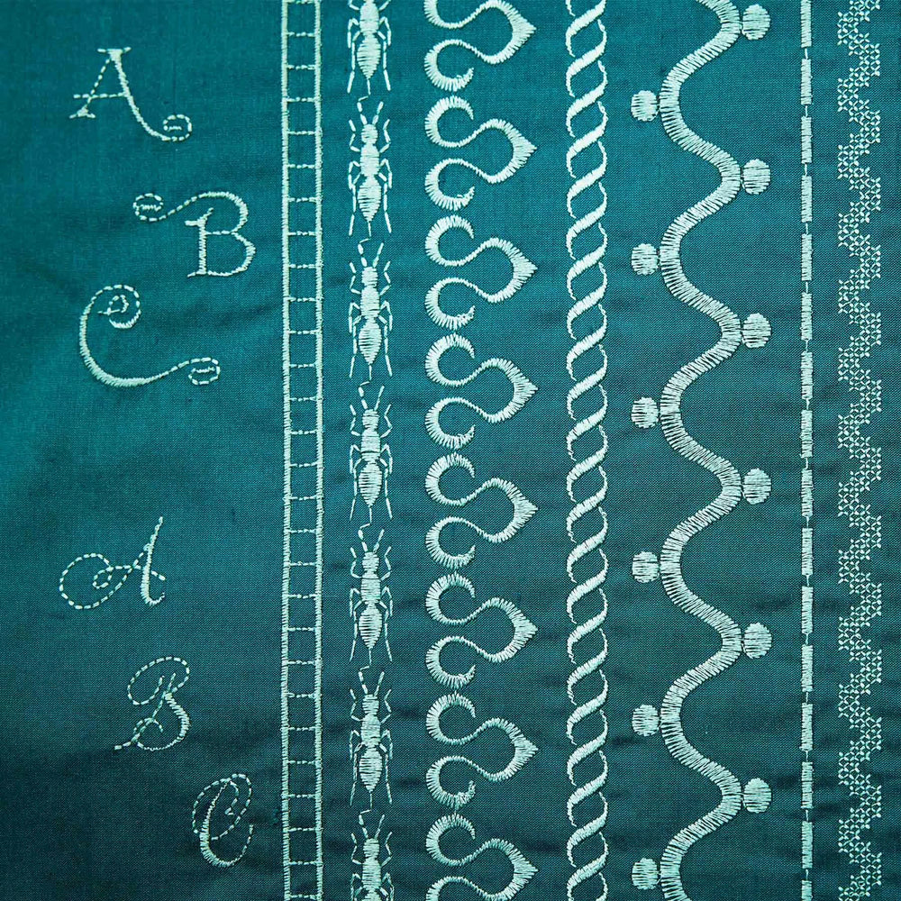 Teal fabric with white embroidery patterns including letters, leaves, and abstract designs.