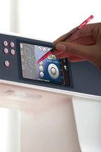 Hand using a red pen to interact with a sewing machine's digital display.