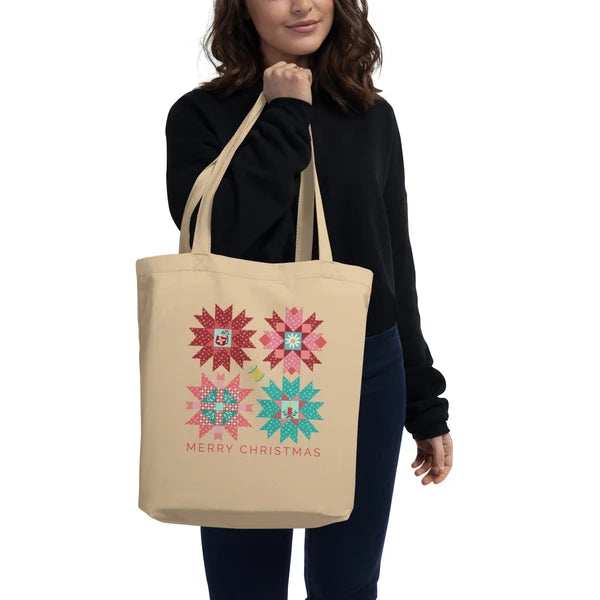 Model carrying the Cozy Cheer tote with four red, pink and teal quilt stars above Merry Christmas lettering.