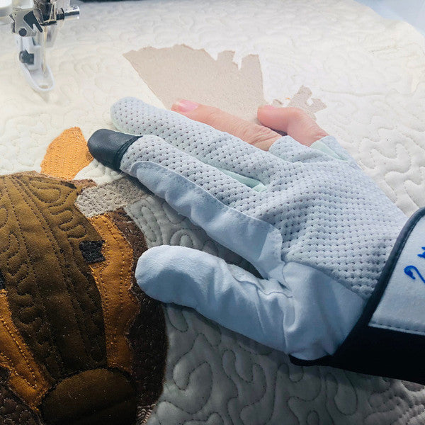 Hands wearing Swan Amity quilting gloves guide an appliqué quilt under a sewing machine, with exposed fingertips visible.