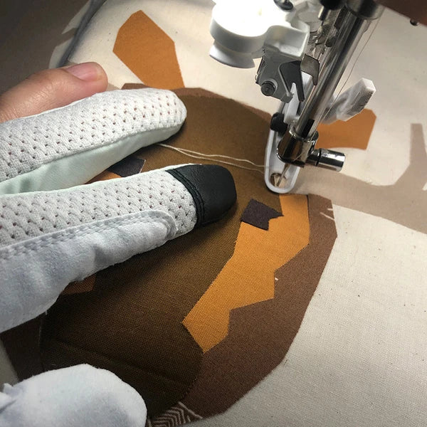 Close-up of Swan Amity gloved fingers guiding brown-and-gold appliqué fabric beside the sewing machine needle.