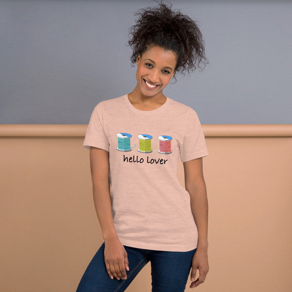 Model wearing the Heather Prism Peach Hello Lover T-shirt with three colorful thread spools above black script lettering.