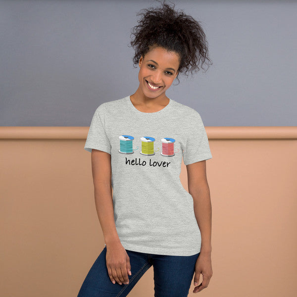 Model wearing the Athletic Heather Hello Lover T-shirt with three colorful thread spools above black script lettering.