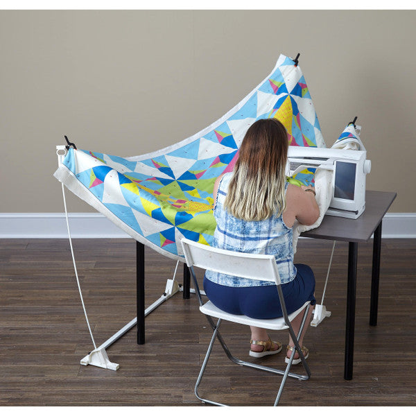 Quilter seated at a sewing machine while the Weightless Quilter frame supports a colorful geometric quilt around the table.